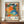 May include: Framed exhibition poster featuring vibrant abstract artwork with colorful dots, flowers, and organic shapes in orange, blue, green, pink, and black. Displayed on wooden credenza beside white ceramic vase and books against cream walls with white trim molding.