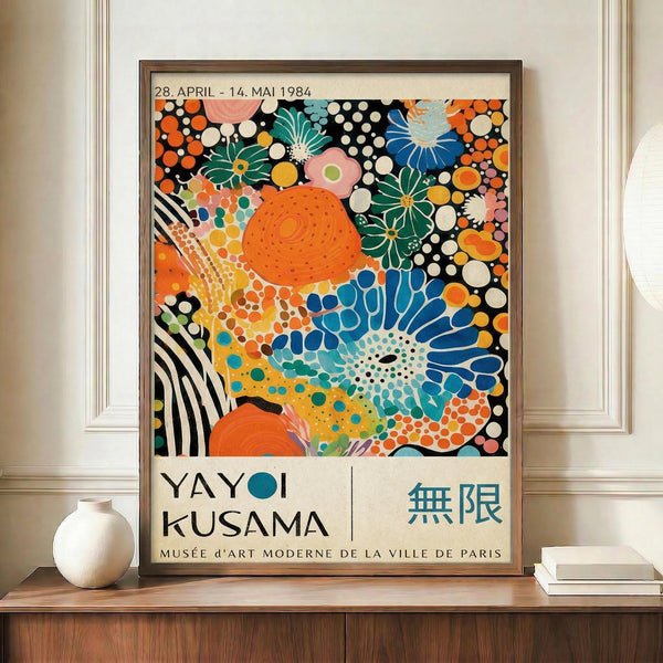 May include: Framed exhibition poster featuring vibrant abstract artwork with colorful dots, flowers, and organic shapes in orange, blue, green, pink, and black. Displayed on wooden credenza beside white ceramic vase and books against cream walls with white trim molding.