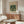 May include: Framed William Morris botanical poster with green parrots and floral pattern displayed above wooden sideboard in modern living room. Cream textured chair, glass oval coffee table, and white table lamp visible. Warm natural lighting creates soft shadows on neutral walls.