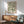 May include: Framed botanical poster with flowing green leaves in swirling pattern on cream background, displayed above wooden sideboard with ceramic table lamp. Mid-century modern sage green upholstered armchair with wooden frame sits nearby. Natural sunlight creates shadows on white wall in minimalist living room setting.