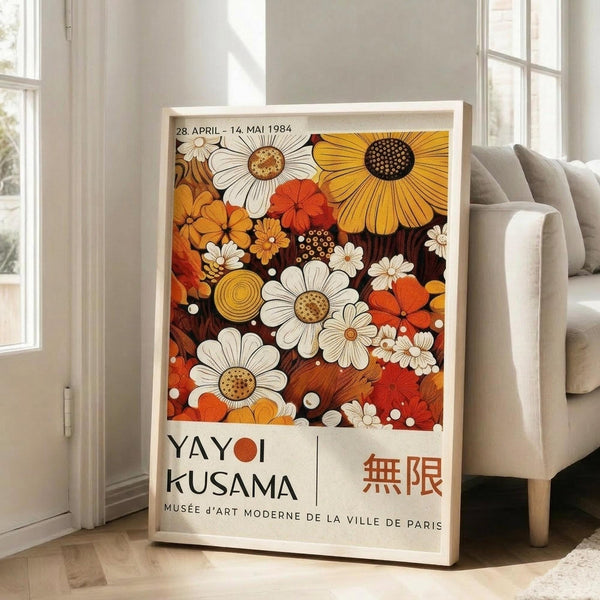 May include: Framed vintage exhibition poster featuring colorful floral pattern with white daisies, orange and yellow blooms on dark background, displayed against white wall next to beige sofa in bright living room with large windows and natural lighting.