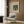 May include: Beige armchair with geometric pillow in modern room setting with wood paneled wall. Framed botanical poster featuring green William Morris pattern with two parrots among leafy vines and flowers hangs above chair. Gold floor lamp and dark round side table with open book complete the warm, neutral-toned interior scene.