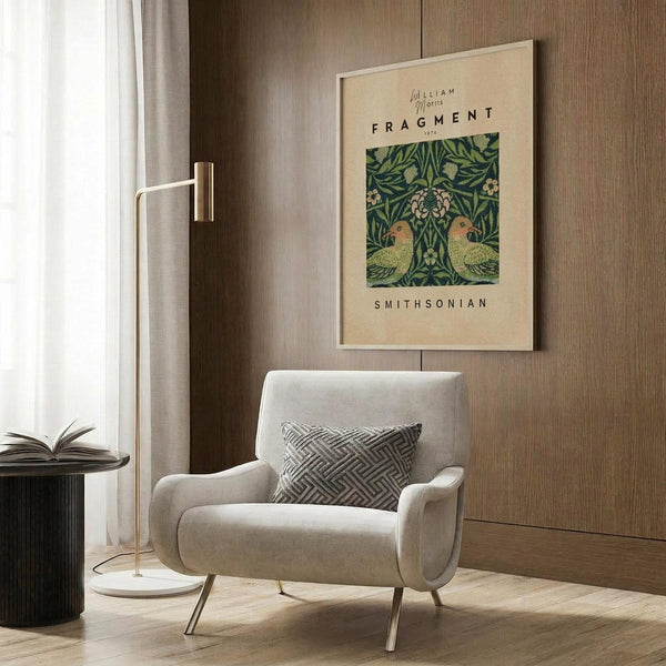 May include: Beige armchair with geometric pillow in modern room setting with wood paneled wall. Framed botanical poster featuring green William Morris pattern with two parrots among leafy vines and flowers hangs above chair. Gold floor lamp and dark round side table with open book complete the warm, neutral-toned interior scene.