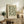 May include: Framed botanical poster with sage green and cream floral pattern leaning against white wall on rustic wood sideboard. Room setting shows fern plant, ceramic vase, stacked books, and beige upholstered chair with brown throw blanket. Soft natural lighting creates warm, cozy atmosphere in modern farmhouse style interior.