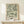 May include: Framed botanical art print with flowing green and cream leaf patterns in wooden frame, displayed on light wood console table beside small wrapped gift with natural twine and greenery sprig, against cream wall with soft natural lighting