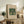 May include: Framed vintage botanical poster featuring two green parrots surrounded by ornate foliage and flowers in William Morris style, displayed on rustic wooden sideboard alongside fern plant, ceramic vase, and stacked books in warm neutral living room setting