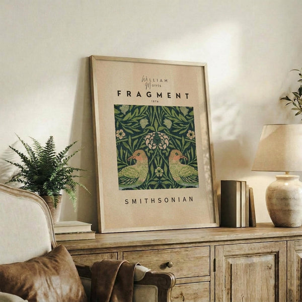 May include: Framed vintage botanical poster featuring two green parrots surrounded by ornate foliage and flowers in William Morris style, displayed on rustic wooden sideboard alongside fern plant, ceramic vase, and stacked books in warm neutral living room setting