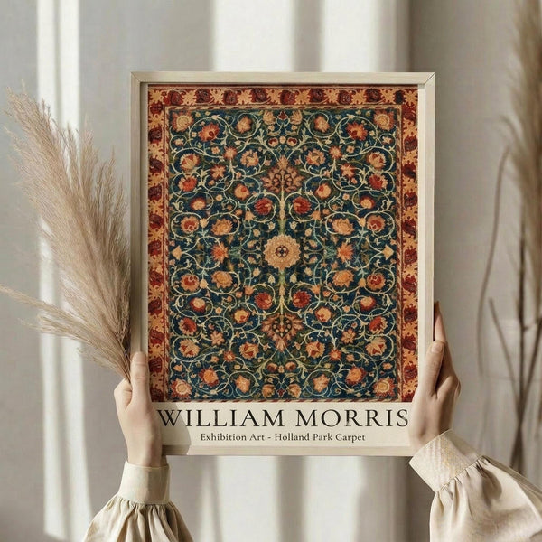 May include: Person in cream clothing holding framed botanical print featuring intricate floral carpet pattern with teal blue center, ornate red and gold border, and detailed flowers throughout. Light wood frame against neutral background with dried wheat stems visible to left side.
