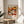 May include: Framed exhibition poster featuring vibrant floral pattern in warm oranges, yellows, and whites with daisies and abstract blooms on dark background. Text shows museum details and Japanese characters. Poster sits on round wooden side table next to cream upholstered chair with plant shadows cast on white wall behind.