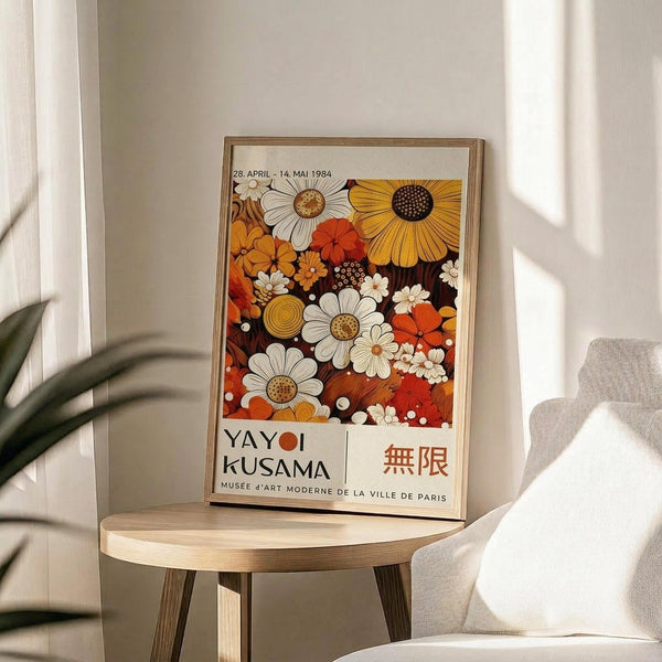 May include: Framed exhibition poster featuring vibrant floral pattern in warm oranges, yellows, and whites with daisies and abstract blooms on dark background. Text shows museum details and Japanese characters. Poster sits on round wooden side table next to cream upholstered chair with plant shadows cast on white wall behind.