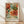 May include: Framed botanical art print leaning against white wall on light wood floor. Artwork features ornate orange and coral peonies with flowing teal and green foliage in decorative Victorian style. Natural wood frame with light finish. Soft natural lighting creates diagonal shadows across white textured wall. Print displays "WILLIAM MORRIS" and "Exhibition Art - Tranquil Botanics" text below floral design.