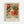 A framed botanical art print featuring ornate floral designs with large blooming flowers in warm coral, orange and red tones alongside decorative leaves in sage green and teal. The vintage-inspired artwork has flowing, ornamental patterns with layered petals and curving foliage against a cream background. Below the botanical illustration is text reading "WILLIAM MORRIS" and "Exhibition Art - Tranquil Botanics" in dark lettering. The print is displayed in a light wood frame against a whit