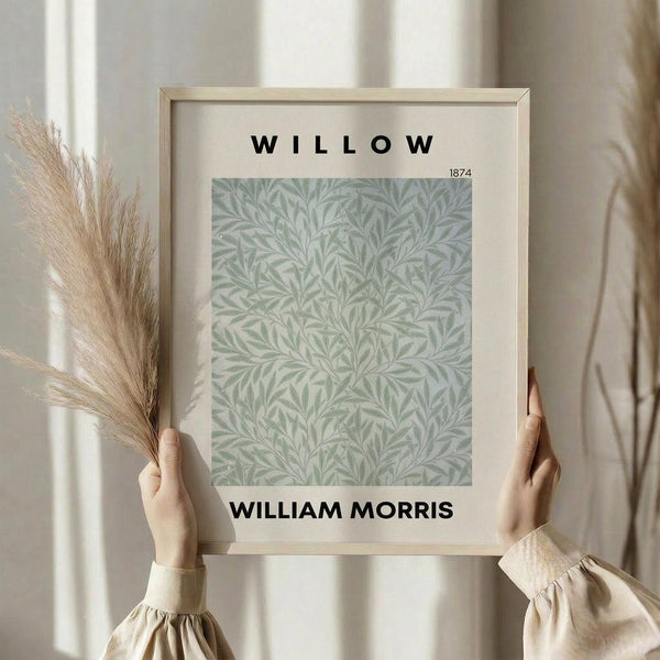 May include: Person in beige ruffled sleeves holding framed botanical print with sage green willow leaf pattern on cream background, natural light streaming through curtains, dried pampas grass stems visible, soft neutral home decor setting with wooden frame.