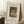 May include: Framed art print leaning against white wall on textured cream rug. Features woman in wide-brimmed hat and green dress holding wicker basket of eggs, surrounded by lush green foliage. Museum-style poster format with cream matting, black text reading "Woman with Basket Eggs" and attribution to William Morris, Barnes Foundation. Small wooden block with white coffee cup visible on left side of scene.