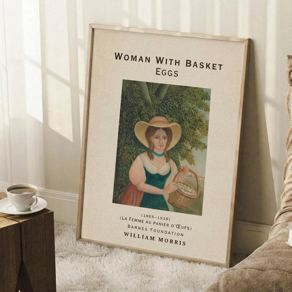 May include: Framed art print leaning against white wall on textured cream rug. Features woman in wide-brimmed hat and green dress holding wicker basket of eggs, surrounded by lush green foliage. Museum-style poster format with cream matting, black text reading "Woman with Basket Eggs" and attribution to William Morris, Barnes Foundation. Small wooden block with white coffee cup visible on left side of scene.