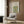 May include: Vintage botanical poster with ornate blue and orange floral vine pattern displayed on wood paneled wall above modern beige armchair with geometric pillow. Gold floor lamp and dark side table with open book complete the neutral-toned reading nook setup. The framed print shows intricate Arts and Crafts style wallpaper design with repeating flower motifs.
