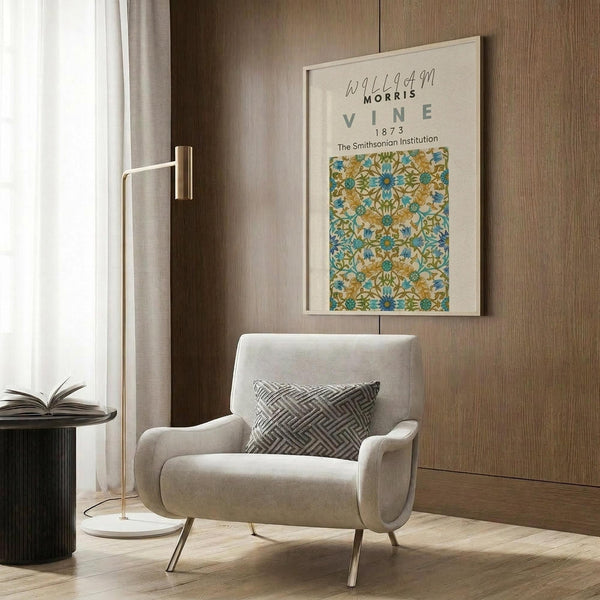 May include: Vintage botanical poster with ornate blue and orange floral vine pattern displayed on wood paneled wall above modern beige armchair with geometric pillow. Gold floor lamp and dark side table with open book complete the neutral-toned reading nook setup. The framed print shows intricate Arts and Crafts style wallpaper design with repeating flower motifs.