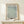 May include: Framed botanical poster with sage green willow leaf pattern on cream background, displayed on light wood console table with small wrapped gift box beside it, against neutral wall in minimalist home setting