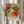 May include: Hands holding a framed botanical poster featuring vibrant orange and coral peonies with teal and green foliage in William Morris decorative style. Light wood frame against neutral curtained background with dried pampas grass visible to the left. Person wearing cream ruffle-sleeved top displaying the colorful floral artwork.