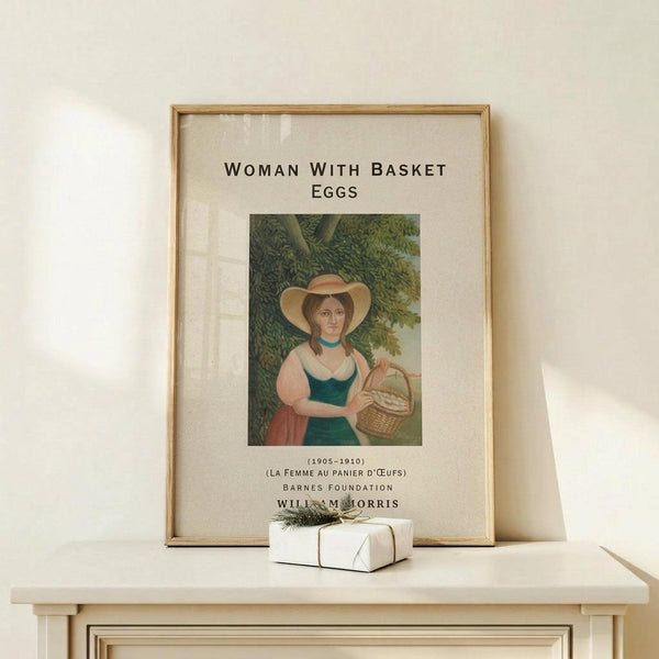 May include: Framed art print displayed on light wood console table against white wall. Print shows woman in wide-brimmed hat and green dress holding wicker basket, surrounded by lush foliage. Natural wood frame with glass front, cream matting. Small wrapped gift with twine and greenery sits on table below. Soft natural lighting creates gentle shadows on minimalist styled surface.