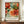 May include: Framed botanical art poster with vibrant orange and coral flowers, teal and green foliage in decorative swirling patterns against cream background. White ceramic vase and stacked books sit beside the wooden-framed artwork on a walnut credenza. Neutral wall with white wainscoting visible in background.