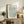 May include: Framed botanical poster with sage green willow leaf pattern displayed on rustic wooden sideboard alongside fern plant, books, and ceramic vase in neutral-toned living room with cream armchair and soft natural lighting