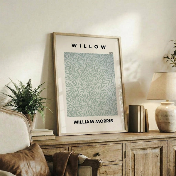 May include: Framed botanical poster with sage green willow leaf pattern displayed on rustic wooden sideboard alongside fern plant, books, and ceramic vase in neutral-toned living room with cream armchair and soft natural lighting