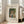 May include: Framed art print leaning against white wall next to beige upholstered chair in bright living room. Print shows woman in green dress and wide-brimmed hat holding wicker basket, surrounded by lush green foliage. Natural wood frame with cream matting. Text reads "WOMAN WITH BASKET EGGS" and includes dates 1905-1910, French title, Barnes Foundation, and William Morris attribution. Sunlight streams through tall windows onto light wooden floors.