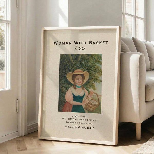 May include: Framed art print leaning against white wall next to beige upholstered chair in bright living room. Print shows woman in green dress and wide-brimmed hat holding wicker basket, surrounded by lush green foliage. Natural wood frame with cream matting. Text reads "WOMAN WITH BASKET EGGS" and includes dates 1905-1910, French title, Barnes Foundation, and William Morris attribution. Sunlight streams through tall windows onto light wooden floors.