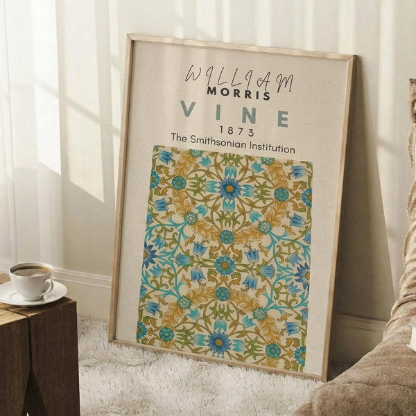 May include: Framed art print featuring William Morris floral vine pattern in blue, teal, and golden yellow colors from 1873, displayed leaning against white wall corner near wooden side table with coffee cup and cozy throw blanket in bright natural lighting