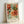 May include: Framed botanical art print featuring ornate orange and coral flowers with flowing turquoise and green foliage in decorative Victorian style, displayed in light wood frame against white wall corner. The artwork shows intricate floral patterns with layered petals and swirling leaves in warm autumn tones.