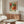 May include: Framed botanical poster with vibrant orange and coral flowers, green foliage on cream background, displayed above walnut wood sideboard in modern living room. White curved chair and glass oval coffee table visible. Natural lighting creates warm shadows on neutral walls.