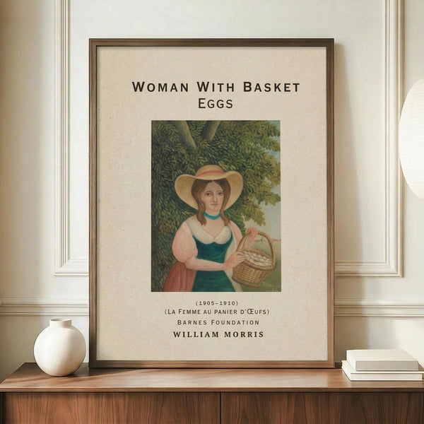May include: Framed vintage-style poster featuring a woman in traditional dress with wide-brimmed hat holding a wicker basket of eggs beneath leafy trees. The artwork has muted green, pink and cream tones with museum-style text labeling. Brown wooden frame displayed on wooden console beside white ceramic vase and stacked books against cream wall with white molding.
