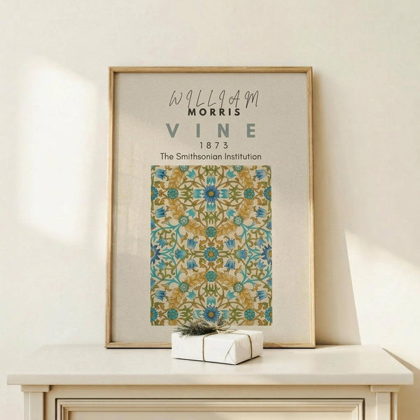 May include: Framed botanical poster with ornate blue and gold floral pattern displayed on light wood console table against cream wall. Small white wrapped gift with evergreen sprig sits in front of natural wood frame. Arts and crafts style wallpaper design features intricate vines, flowers and decorative motifs in teal, golden yellow and green tones.