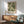 May include: Framed botanical print with flowing green and cream leaf pattern hanging above wooden sideboard. Mid-century modern room setting with sage green upholstered chair, white ceramic table lamp, and natural sunlight casting shadows on white wall. Print features swirling acanthus leaves in muted earth tones with exhibition-style text below artwork.