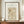 May include: Framed poster displaying William Morris Garden Tulip pattern from 1885 Birmingham, featuring red tulip flowers on green stems against an ornate sage green and cream damask background. The print sits on a wooden sideboard beside a small white ceramic vase and stacked books in a neutral-toned room with white wainscoting.