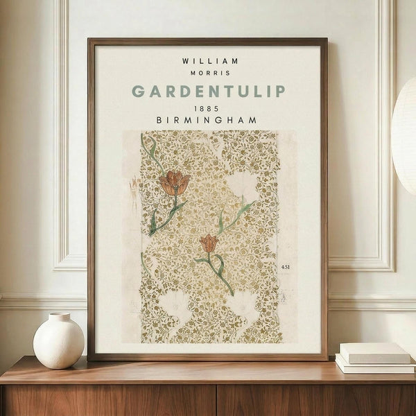 May include: Framed poster displaying William Morris Garden Tulip pattern from 1885 Birmingham, featuring red tulip flowers on green stems against an ornate sage green and cream damask background. The print sits on a wooden sideboard beside a small white ceramic vase and stacked books in a neutral-toned room with white wainscoting.