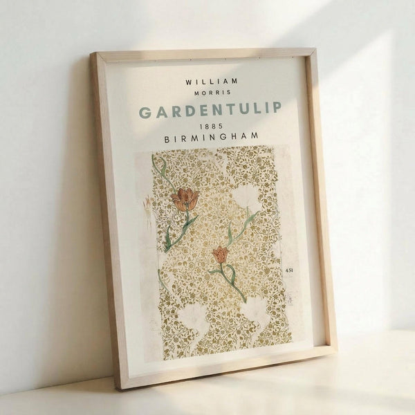 May include: Framed vintage botanical print featuring William Morris Garden Tulip design from 1885 Birmingham. The artwork shows delicate orange-red tulip flowers with green stems against an intricate golden-brown damask pattern background. Natural wood frame displayed at an angle against white wall, showing warm lighting and shadows.