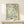May include: Framed botanical art print with flowing green foliage pattern on cream background, displayed on light wood console table against white wall. Small white gift box with greenery sprig sits in front of the wooden-framed artwork. Soft natural lighting creates gentle shadows on the minimalist styled surface.