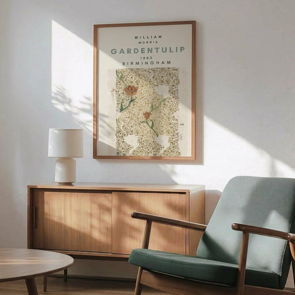 May include: Framed botanical print featuring William Morris Garden Tulip design from 1885 Birmingham, displayed above a wooden sideboard with table lamp. Mid-century modern sage green armchair with wooden arms sits beside a round coffee table. Warm sunlight creates shadows on white wall, showcasing the vintage floral pattern in muted greens and browns within a thin wooden frame.