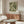 May include: Framed botanical poster with flowing green and cream acanthus leaf pattern hanging above walnut credenza in minimalist living room. White curved chair and glass coffee table visible in foreground. Natural lighting creates soft shadows on beige walls.