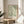 May include: Framed poster with flowing green botanical design featuring swirling leaves and foliage on cream background, displayed on round wooden side table next to beige upholstered chair. Natural sunlight creates soft shadows across white wall. Live plant leaves visible in corner of minimalist, neutral-toned room setting.