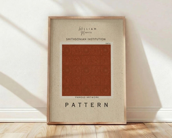 May include: Framed poster print leaning against white wall on light wooden floor. Features cream-colored background with rust orange geometric pattern square in center. Text reads "William Morris," "Smithsonian Institution 1870," "Famous Artwork," and "PATTERN" in black typography. Natural wood frame with warm lighting casting diagonal shadows across the scene.