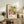 May include: Colorful vintage Yayoi Kusama exhibition poster with vibrant polka-dotted circular patterns in pink, yellow, green, and orange, displayed in light wood frame on rustic wooden sideboard. Room scene shows beige armchair, fern plant in white pot, ceramic vases, and stacked books creating warm, modern living space with neutral wall.