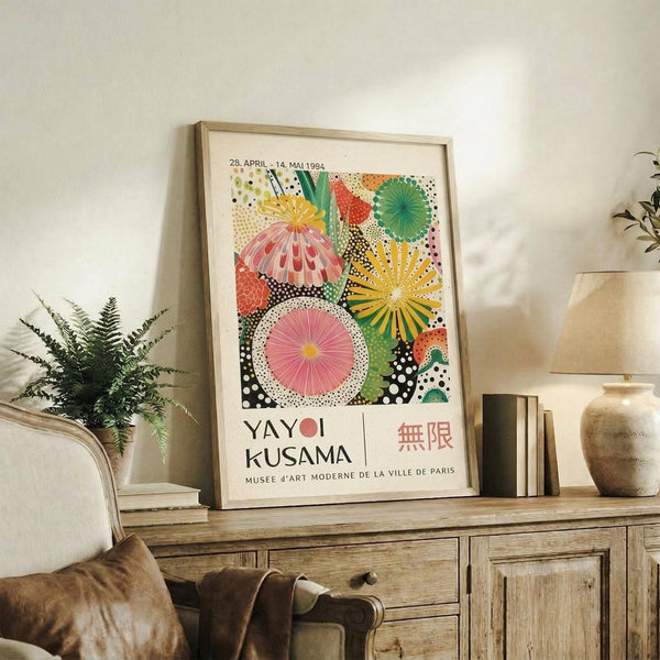 May include: Colorful vintage Yayoi Kusama exhibition poster with vibrant polka-dotted circular patterns in pink, yellow, green, and orange, displayed in light wood frame on rustic wooden sideboard. Room scene shows beige armchair, fern plant in white pot, ceramic vases, and stacked books creating warm, modern living space with neutral wall.