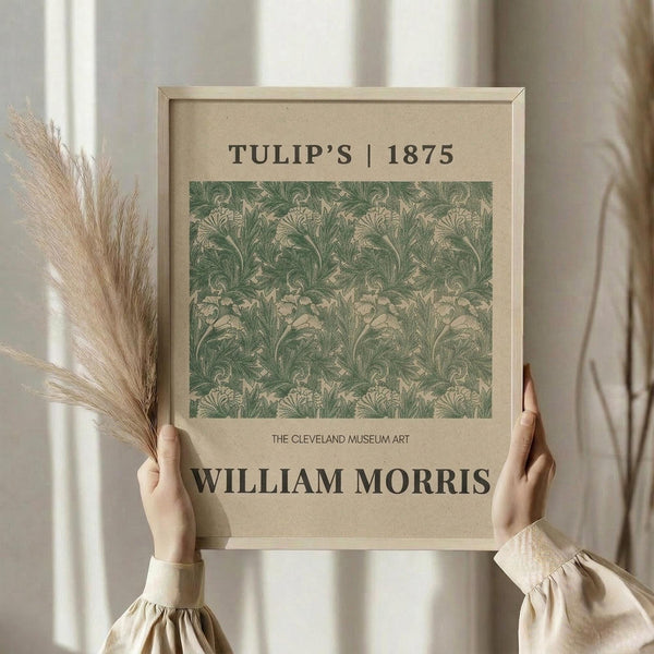 May include: Hands holding a framed vintage botanical print featuring an intricate green and cream floral pattern with tulip motifs in an ornate Victorian style. The artwork is displayed in a light wood frame against soft neutral curtains, with dried pampas grass visible to the side, creating a minimalist aesthetic room setting.