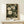 May include: Framed botanical poster with cream-colored flowers and dark green foliage on black background, displayed on light wood console table with small white gift box, against neutral wall in minimalist interior setting