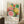 May include: Framed vintage exhibition poster featuring colorful abstract floral and circular patterns in pink, yellow, green, and orange with polka dot backgrounds. Shows dates "28. APRIL - 14. MAI 1984" and "YAYOI KUSAMA" text with "MUSÉE D'ART MODERNE DE LA VILLE DE PARIS" below. Japanese characters visible in corner. Poster leans against white wall next to small wooden side table with white coffee cup and saucer, positioned on white textured rug with beige throw blanket