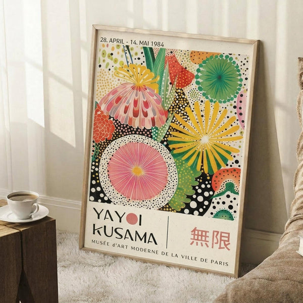 May include: Framed vintage exhibition poster featuring colorful abstract floral and circular patterns in pink, yellow, green, and orange with polka dot backgrounds. Shows dates "28. APRIL - 14. MAI 1984" and "YAYOI KUSAMA" text with "MUSÉE D'ART MODERNE DE LA VILLE DE PARIS" below. Japanese characters visible in corner. Poster leans against white wall next to small wooden side table with white coffee cup and saucer, positioned on white textured rug with beige throw blanket