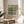 May include: Framed vintage botanical poster with green and cream tulip pattern design displayed on round wooden side table next to beige upholstered chair. Natural sunlight streams across white walls, with plant leaves visible in corner. Poster shows decorative Arts and Crafts style floral motif in sage green tones with museum exhibition text.